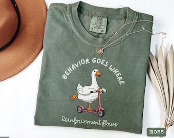 May include: Moss green t-shirt with a graphic of a white goose riding a pink scooter. The text "BEHAVIOR GOES WHERE" and "Reinforcement flows" are printed on the shirt. A gold necklace and a brown hat are also visible.
