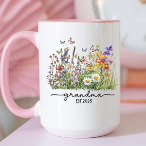 May include: White ceramic mug with a pink handle and rim, decorated with a colorful floral design and butterflies. The word "grandma" and the year "EST. 2025" are written in a script font. A charming gift idea.