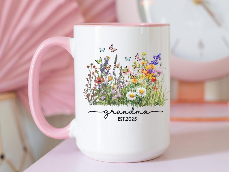 May include: White ceramic mug with a pink handle and rim, decorated with a colorful floral design and butterflies. The word "grandma" and the year "EST. 2025" are written in a script font. A charming gift idea.