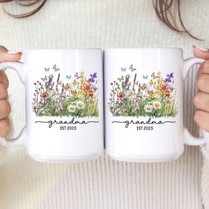 May include: Two white ceramic mugs with a floral design featuring colorful wildflowers and butterflies. The word "grandma" is written in script, with "EST.2025" below. The mugs are held by a person.