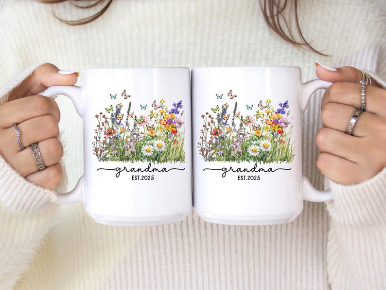 May include: Two white ceramic mugs with a floral design featuring colorful wildflowers and butterflies. The word "grandma" is written in script, with "EST.2025" below. The mugs are held by a person.