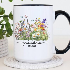 May include: White ceramic mug with a black handle and rim, featuring a floral design with butterflies. The mug has the word "grandma" in script, with "EST.2025" below the word.