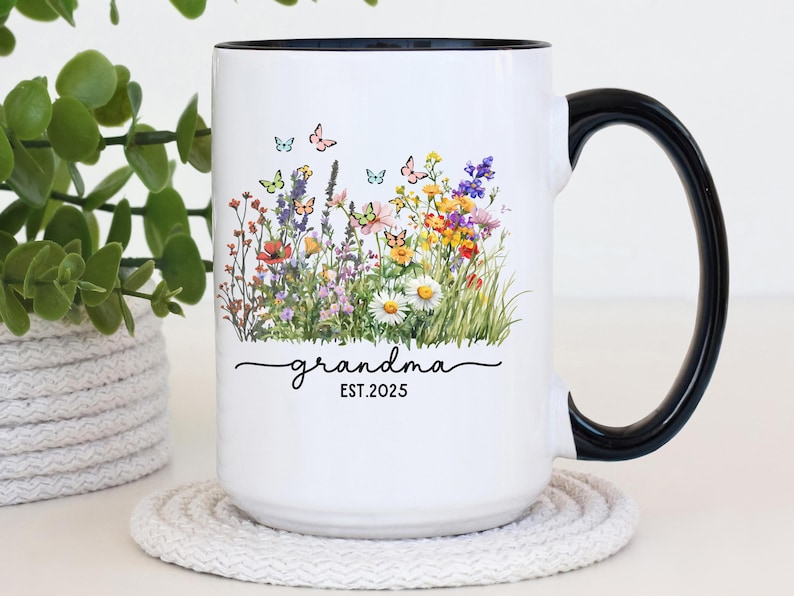 May include: White ceramic mug with a black handle and rim, featuring a floral design with butterflies. The mug has the word "grandma" in script, with "EST.2025" below the word.