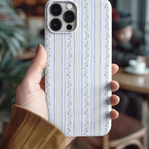 May include: A white phone case with a floral and striped pattern. The case features vertical stripes in light blue and green, with delicate floral accents. The phone case is held in a person's hand.