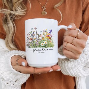 May include: A white ceramic mug with a black handle and the text "But First, Coffee" in a bold, black font. The mug is set against a blurred background, suggesting a cozy setting.