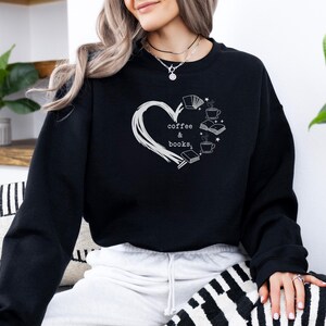 May include: Black sweatshirt with a white heart-shaped design. Inside the heart are the words "coffee & books" and illustrations of books and coffee cups. The sweatshirt is long-sleeved and made of a soft material.