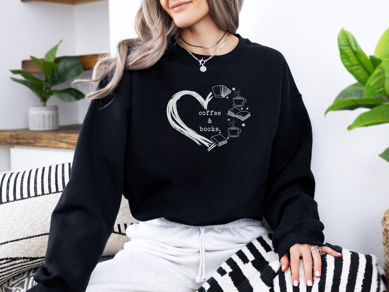 May include: Black sweatshirt with a white heart-shaped design. Inside the heart are the words "coffee & books" and illustrations of books and coffee cups. The sweatshirt is long-sleeved and made of a soft material.