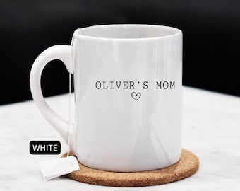 May include: White ceramic mug with the text "OLIVER'S MOM" and a heart symbol. The mug sits on a round cork coaster, with a tea bag attached. The word "WHITE" is in a black rectangle.