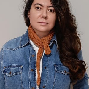 Puede incluir: Una bufanda de punto color óxido se lleva sobre una camisa vaquera. La bufanda es de color liso y parece estar hecha de un hilo suave. La camisa vaquera tiene dos bolsillos delanteros y cierres de botón. El fondo es una pared lisa de color gris claro.