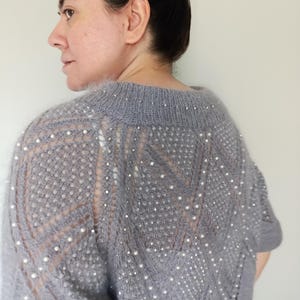 May include: A gray, open-knit sweater with short sleeves and a round neckline. The sweater is embellished with small, round, white beads. The knit pattern features a chevron design on the back and sleeves. The sweater is made of a soft, fuzzy material.
