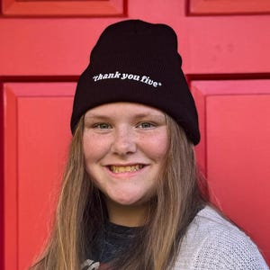 May include: A black beanie with the text "thank you five" in white. The beanie is worn by a person. The person is wearing a gray cardigan and a dark t-shirt. The background is a red door.