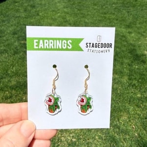 May include: A pair of novelty earrings featuring a cartoon plant design with a red mouth and green leaves. The earrings hang from gold-colored hooks. The earrings are displayed on a white card with the word "EARRINGS" in green.