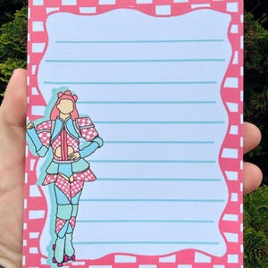 May include: A lined notepad with a pink and white checkered border. The notepad features a cartoon illustration of a person in a roller skating outfit. The outfit is pink, blue, and white.