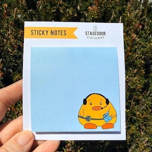 May include: A light blue sticky note pad with a cartoon duck wearing a headset and holding a notepad that says "SCRIBBLE". The pad has the text "STICKY NOTES" and "STAGEDOOR STATIONERY" printed on it.