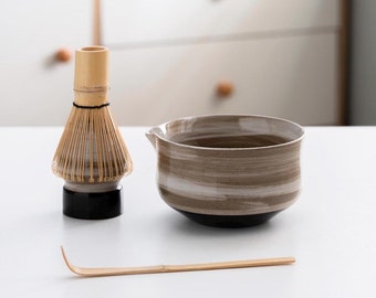 Könnte beinhalten: Eine braune und weiße Keramik-Matcha-Schale mit schwarzem Boden, ein Bambus-Schneebesen mit schwarzem Boden und ein Bambus-Matcha-Löffel auf einer weißen Oberfläche.