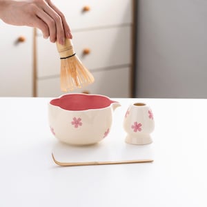 May include: A white ceramic matcha bowl with a pink rim and pink flower designs, a white ceramic matcha whisk holder with pink flower designs, a bamboo matcha whisk, and a bamboo matcha scoop.