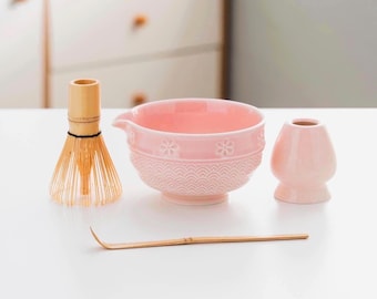 May include: A pink ceramic matcha bowl set with a bamboo whisk, scoop, and a small vase-shaped holder. The bowl has a floral and wave pattern. The whisk is made of thin bamboo tines.