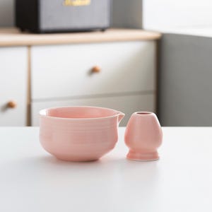 以下が含まれることがあります： ピンクのセラミック製の茶碗と、それに合わせたピンクのセラミック製の茶筅。茶碗は丸みを帯びた形をしていて、小さな注ぎ口が付いている。茶筅は丸みを帯びた取っ手と、長く細い茶筅が付いている。