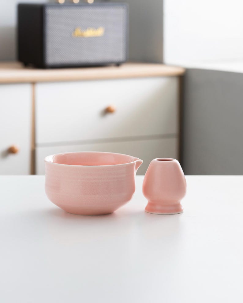以下が含まれることがあります： ピンクのセラミック製の茶碗と、それに合わせたピンクのセラミック製の茶筅。茶碗は丸みを帯びた形をしていて、小さな注ぎ口が付いている。茶筅は丸みを帯びた取っ手と、長く細い茶筅が付いている。