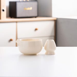 May include: Two off-white ceramic teacups, one with a spout, on a white surface.