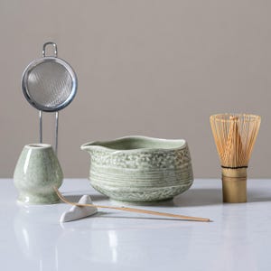 May include: A matcha tea set comprising a light green ceramic bowl, whisk, and holder, a metal sieve, and a bamboo whisk. The set is arranged on a white surface, ideal for preparing traditional matcha tea.