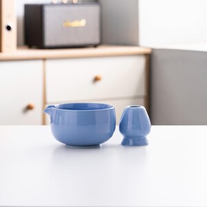 May include: A light blue ceramic teacup and a matching tea whisk on a white surface.