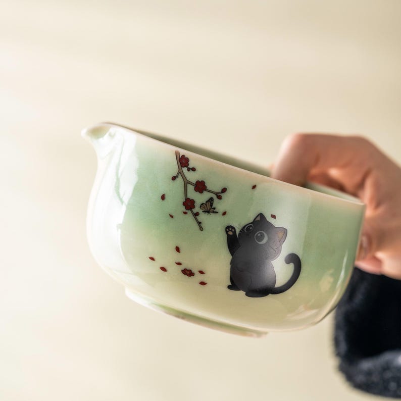May include: A light green ceramic tea pitcher with a black cat illustration. The pitcher has a spout and is decorated with red flower and butterfly accents. The cat is waving its paw.