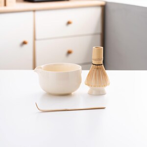May include: A white ceramic tea bowl with a bamboo whisk and a wooden tea scoop on a white surface.