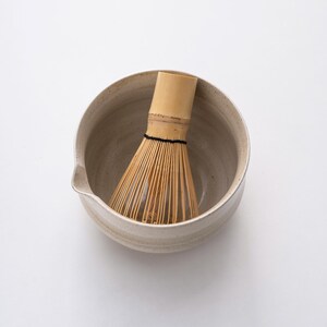 May include: A white ceramic matcha bowl with a bamboo whisk inside. The bowl has a textured surface and a slight lip.