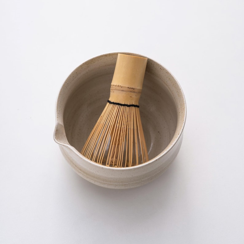 May include: A white ceramic matcha bowl with a bamboo whisk inside. The bowl has a textured surface and a slight lip.