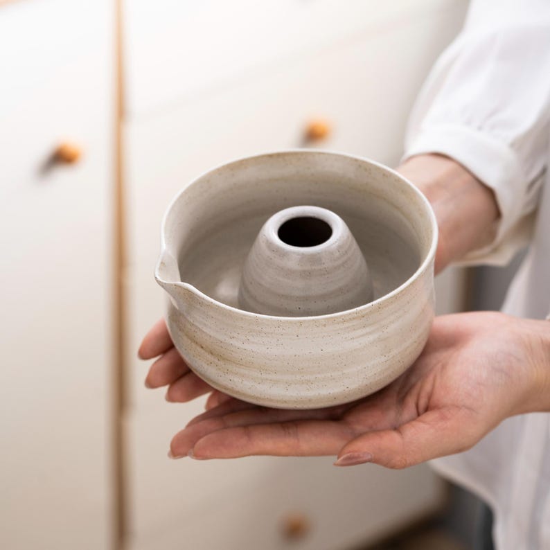 May include: A white ceramic cake mould with a central cone shaped insert. The mould has a speckled glaze and a slightly textured surface.