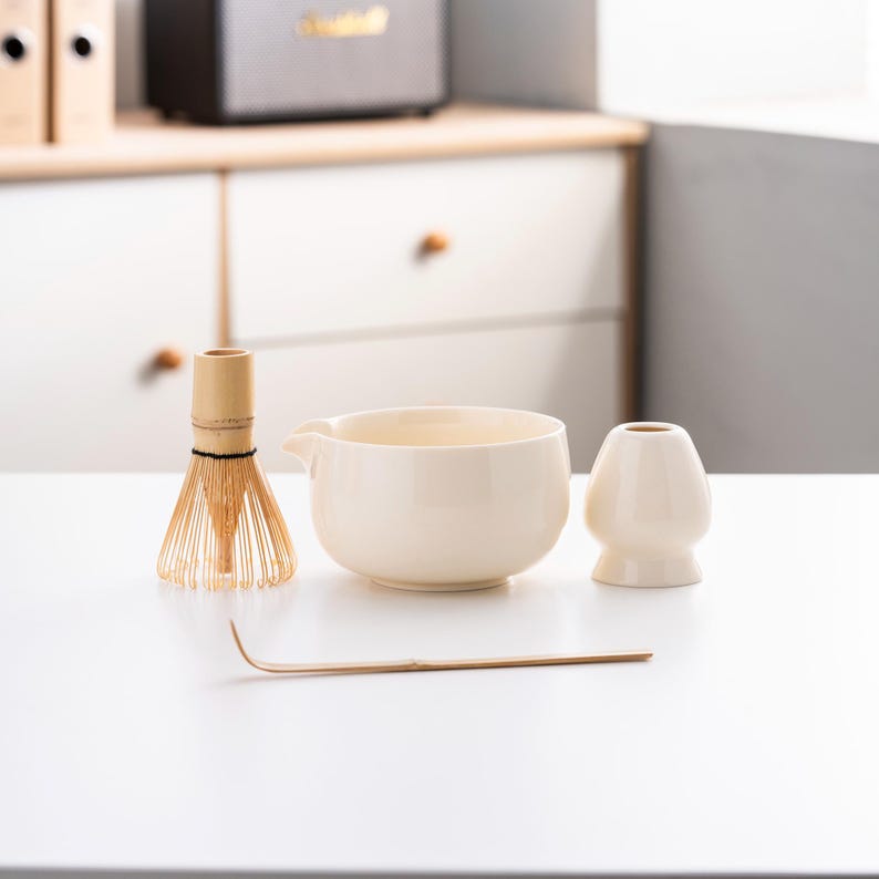 May include: A white ceramic matcha bowl, a bamboo whisk, a white ceramic matcha scoop, and a bamboo matcha whisk holder on a white surface.