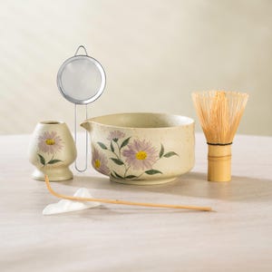 May include: A matcha tea set featuring a ceramic bowl and whisk, a small ceramic container, a metal strainer, and a bamboo whisk holder. The bowl and container are decorated with purple and yellow floral designs.