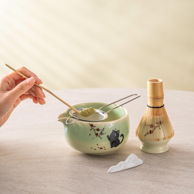 May include: A matcha tea set featuring a light green ceramic bowl with a black cat illustration, a bamboo whisk, a stainless steel strainer, and a bamboo scoop. The bowl has a spout and a white ceramic mountain-shaped rest.