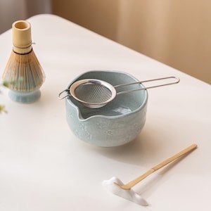 Puede incluir: Un juego de t&eacute; matcha con un cuenco de cer&aacute;mica azul claro con detalles florales, un colador de acero inoxidable, un batidor de bamb&uacute; y una cuchara de bamb&uacute; sobre un soporte de cer&aacute;mica blanca. Para preparar t&eacute; matcha.