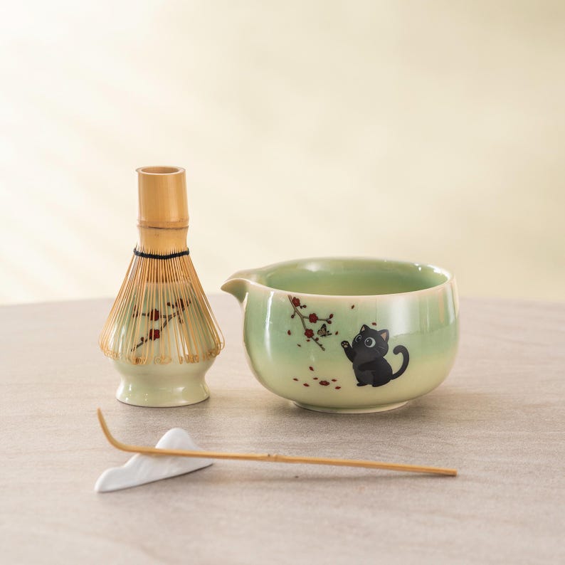 May include: A matcha tea set featuring a light green ceramic bowl with a black cat illustration, a bamboo whisk, and a white ceramic whisk holder with a bamboo whisk. The bowl has a small spout and a floral design.