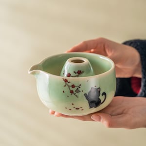 May include: A celadon green ceramic tea set with a black cat illustration and red floral accents. The set includes a bowl with a spout and a central, inverted cone-shaped vessel. The tea set is held in two hands.