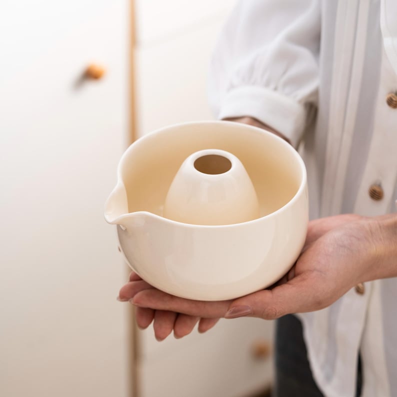May include: A white ceramic pouring pitcher with a central spout and a wide, round base. The pitcher has a unique design with a raised, circular opening in the center.