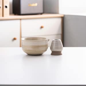 May include: A white ceramic matcha bowl with a brown rim and a white ceramic matcha whisk holder.