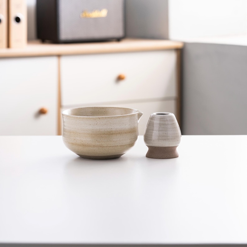 May include: A white ceramic matcha bowl with a brown rim and a white ceramic matcha whisk holder.