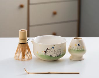 May include: A white ceramic matcha bowl with a cartoon cat design, a bamboo whisk, a bamboo whisk holder, and a white ceramic matcha cup with a green and white floral design.