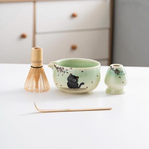 Puede incluir: Un tazón de matcha de cerámica verde con un diseño de gato negro, un batidor de bambú, un soporte para batidor de bambú y una cuchara de matcha de bambú.