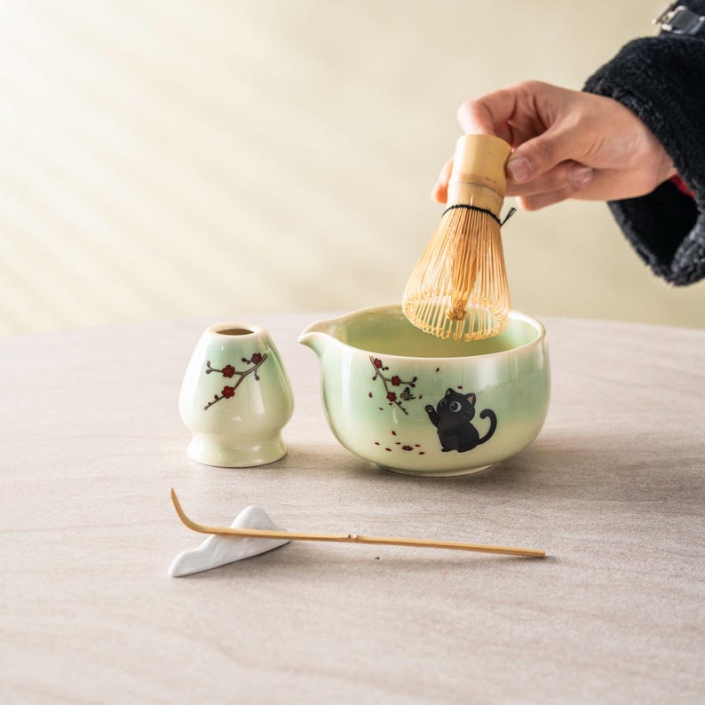 May include: A matcha tea set featuring a light green ceramic bowl with a black cat illustration, a bamboo whisk, a small ceramic holder, and a bamboo scoop. The set includes a small ceramic container with a floral design.