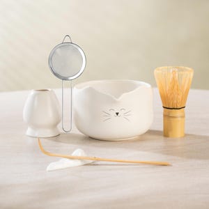 May include: A white ceramic matcha tea set with a cat face design. The set includes a whisk, bowl, whisk holder, strainer, and bamboo scoop. The whisk is made of bamboo and the bowl has a cute cat face.
