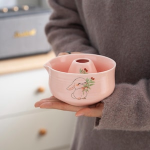 以下が含まれることがあります： ウサギと花柄のピンクのセラミックピッチャー。ピッチャーは注ぎ口とハンドルが付いています。