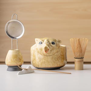 May include: A ceramic matcha set featuring a beige, animal-shaped bowl with a wide open mouth, a matching whisk stand with a metal mesh strainer, a bamboo whisk, and a thin bamboo scoop resting on a white, mountain-shaped ceramic holder.