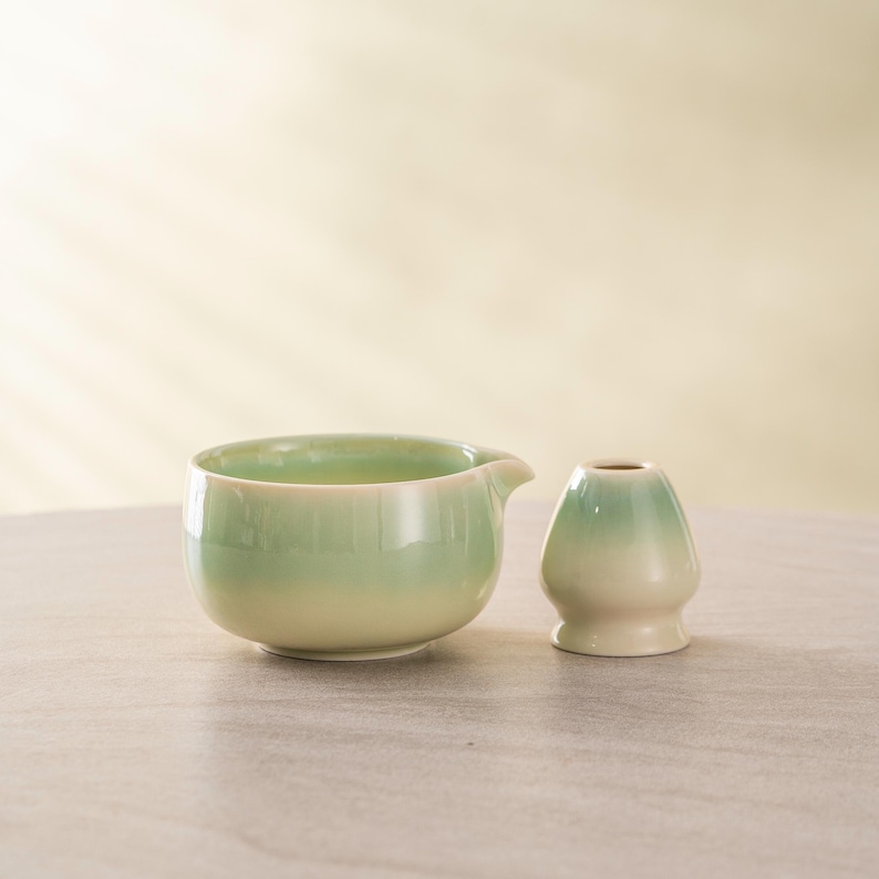 May include: A ceramic tea set featuring a light green and cream-colored bowl with a spout and a matching whisk holder. The set has a glossy finish and is placed on a neutral surface.