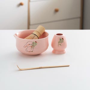 以下が含まれることがあります： ウサギのデザインが施されたピンクのセラミック製の抹茶ボウル、竹製の泡立て器、花柄のピンクのセラミック製の茶碗、竹製の抹茶スプーン