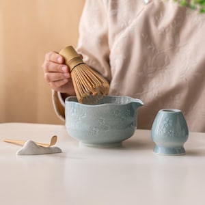Puede incluir: Un juego de t&eacute; matcha de cer&aacute;mica azul claro con motivos florales. El juego incluye un cuenco, un batidor, un soporte para batidor y un jarr&oacute;n peque&ntilde;o. Un batidor de bamb&uacute; se sostiene sobre el cuenco. El juego est&aacute; dise&ntilde;ado para preparar t&eacute; matcha.