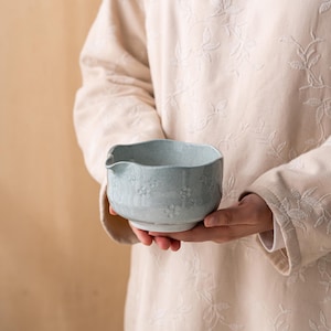 Puede incluir: Un cuenco vertedor de cer&aacute;mica azul claro con un acabado moteado y patrones florales en relieve en el exterior. El cuenco presenta un peque&ntilde;o pico vertedor en un lado y un borde redondeado ligeramente irregular, con una textura esmaltada.